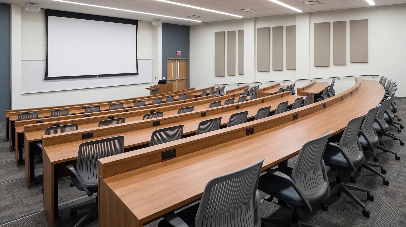 A tiered lecture theatre with long curved wooden desks in rows, grey task chairs on castors, power outlets set into each desk, and a projection screen and lectern at the front.