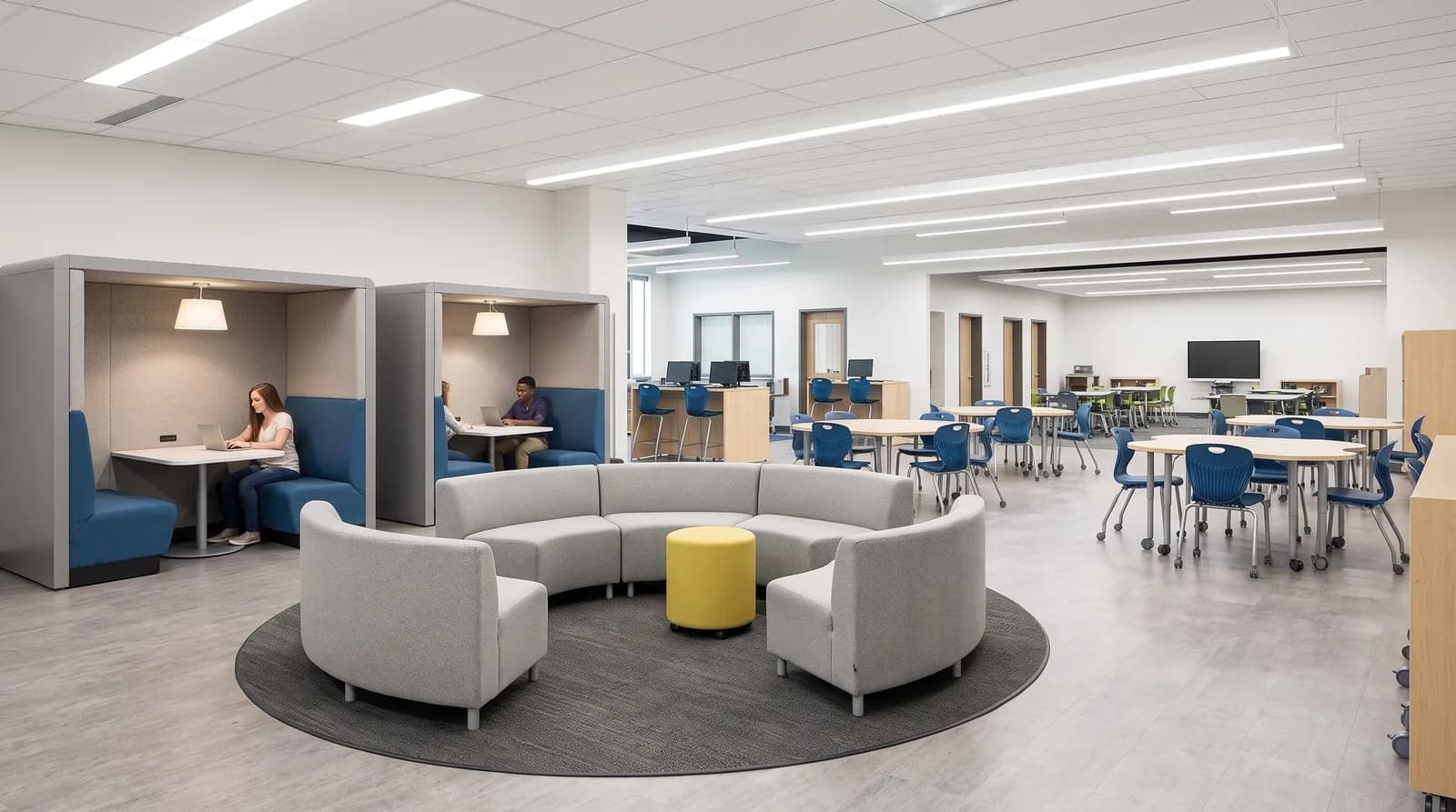 A resource commons where students work at high-backed upholstered study booths with individual lamps, beside a circular grey sofa and yellow ottoman, with round tables and blue chairs in the open area beyond.